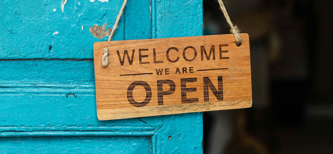 Picture of a sign reading Welcome, we are Open, hanging on a door