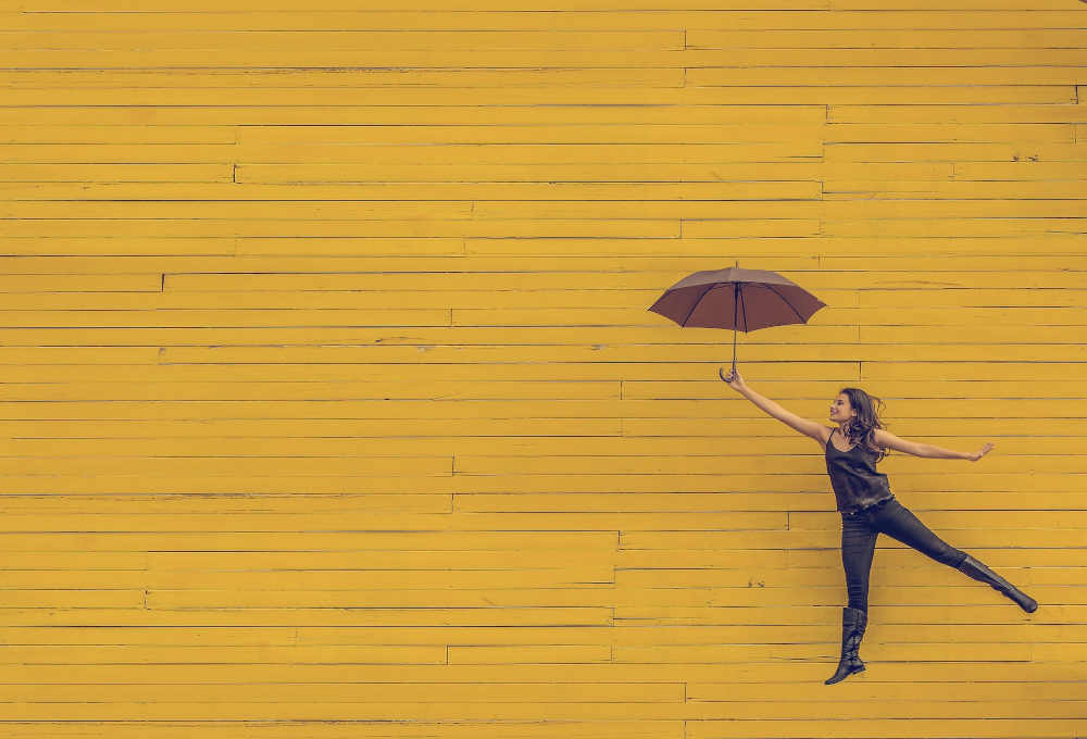 Woman with umbrella jumping in the air in front of a yellow wall.